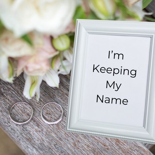 Silver wedding rings and a silver framed
graphic that reads "I'm Keeping My Name"
on weathered gray boards with pastel wedding
flowers in the background
