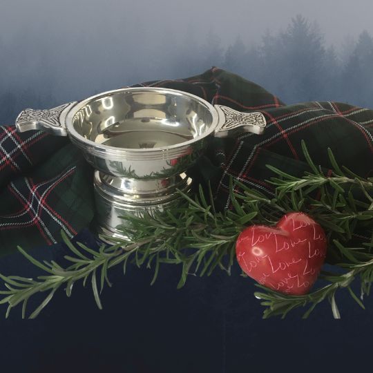 A silver
quaich with a sprig of rosemary, and red
heart-shaped stone that has the word Love repeated
over its surface and a length of the Scottish
National Tartan, on a white background